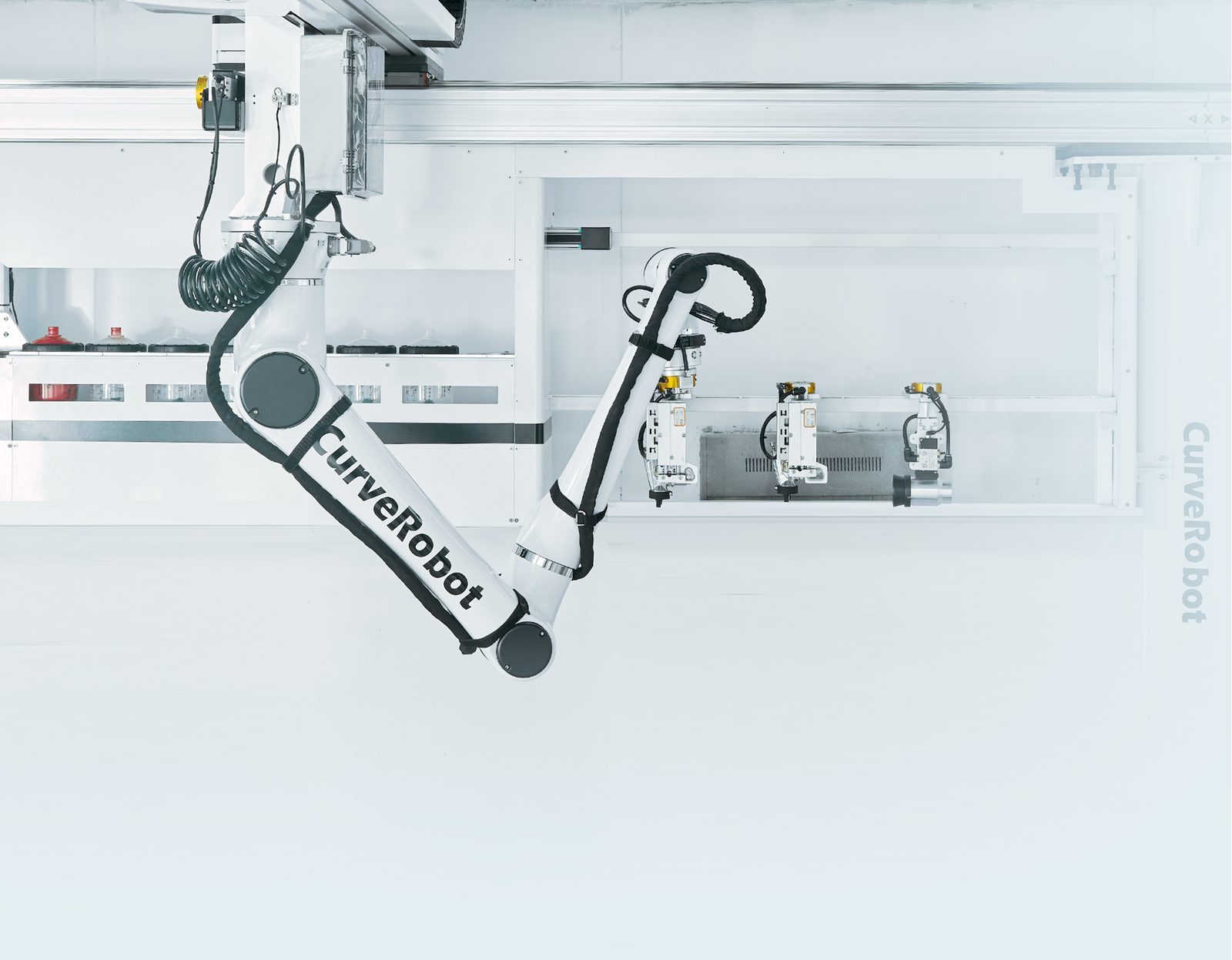 Braccio robotico bianco in cabina, con la rastrelliera delle pistole sulla parete.